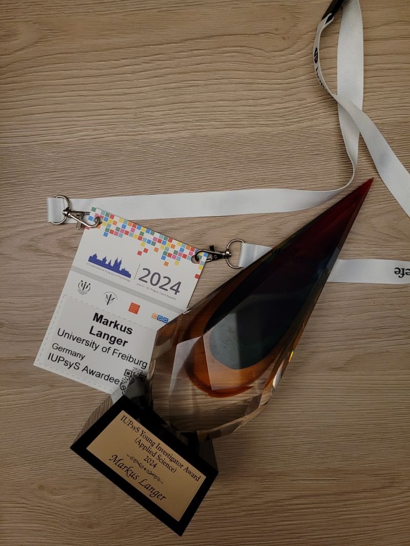 Glastrophäe des IUPsyS Young Investigator Award (Applied Science) 2024 mit Namensschild „Markus Langer“, daneben das Kongress-Namensschild vom International Congress of Psychology in Prag